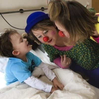 "Hamburger Klinik-Clowns“ in Deutschland, Kind im Krankenhausbett und zwei weibliche Clowns, © Klinik-Clowns Hamburg: Michaela Kuhn