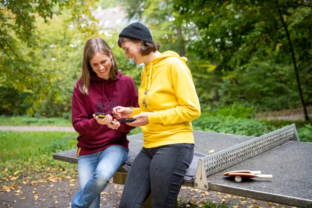 Zwei Frauen stehen vor einer Tischtennisplatte. Sie tragen Pullover von WEtell und schauen gemeinsam auf ein Handy.