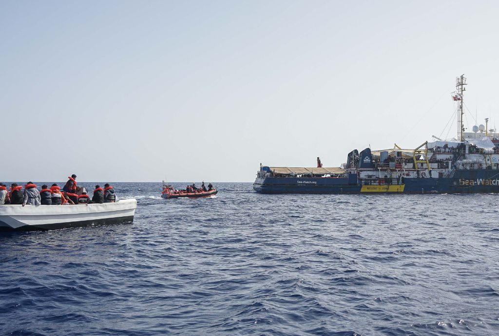 Das Bild zeigt Menschen auf einem Boot im Meer, die von einem Schiff des Vereins Sea-Watch gerettet werden.