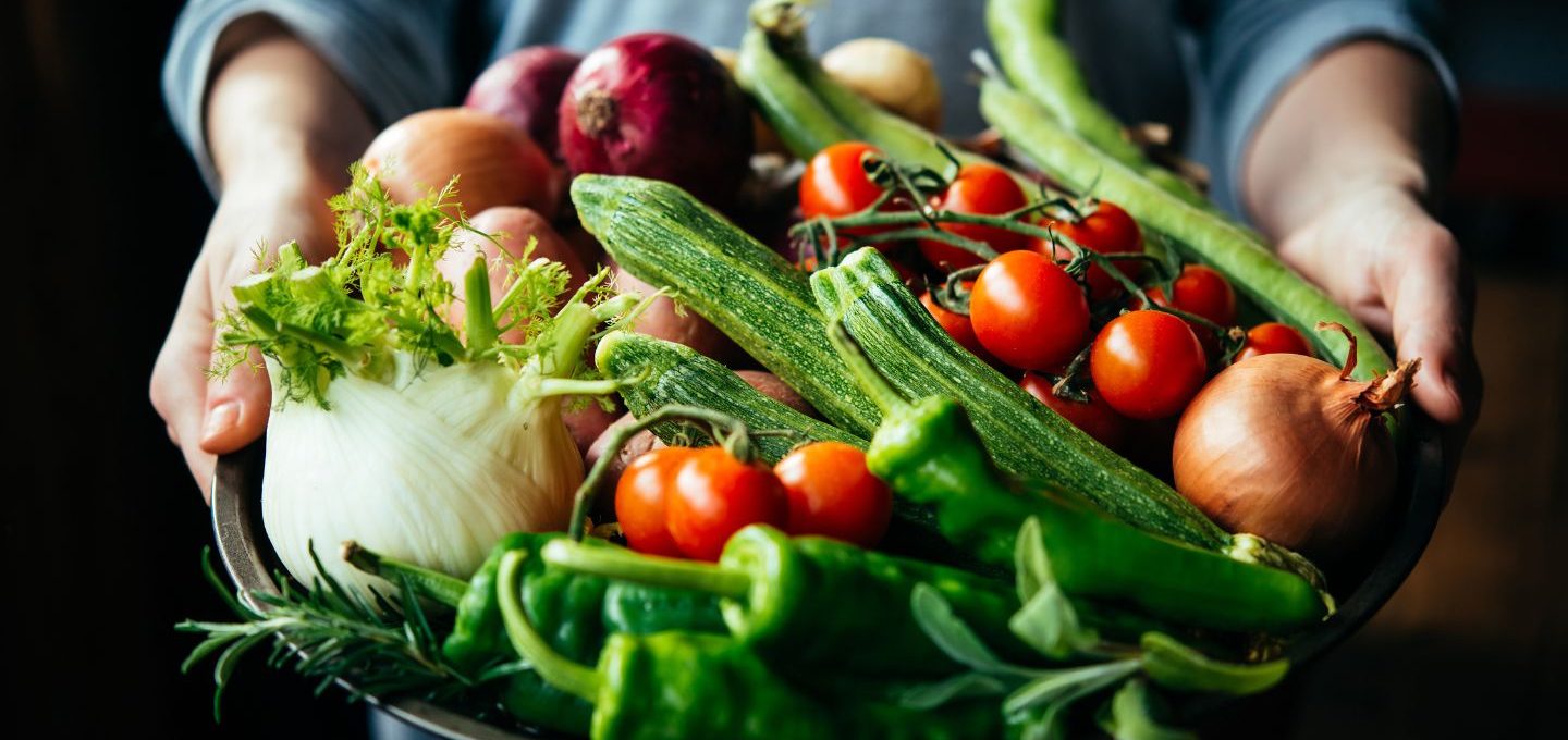 Ein Korb voller Gemüse wird von zwei Händen getragen.