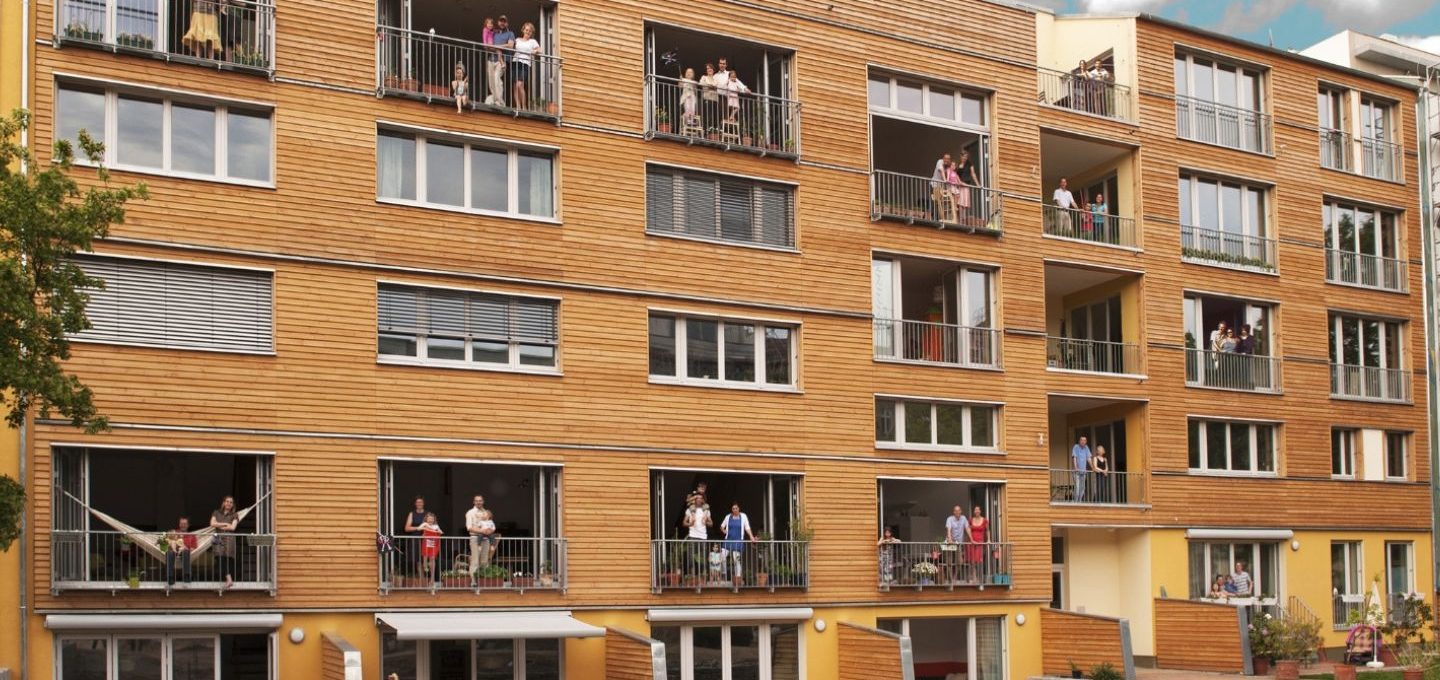Sanieren mit Herz: Das Bild zeigt ein Mehrfamilienhaus in Holzbauweise mit Balkonen, auf denen Menschen stehen.
