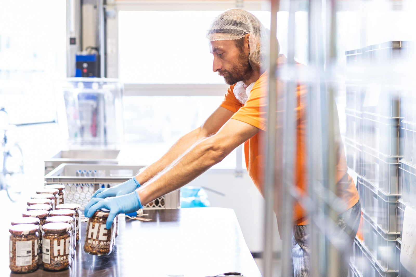 Daniel steht in der Produktionshalle. Er ist von der Seite zu sehen. Er trägt ein oranges T-Shirt und eine Haube über den Haaren und blaue Kunststoff-Handschuhe. Er räumt Müsli-Gläser in eine Stiege.