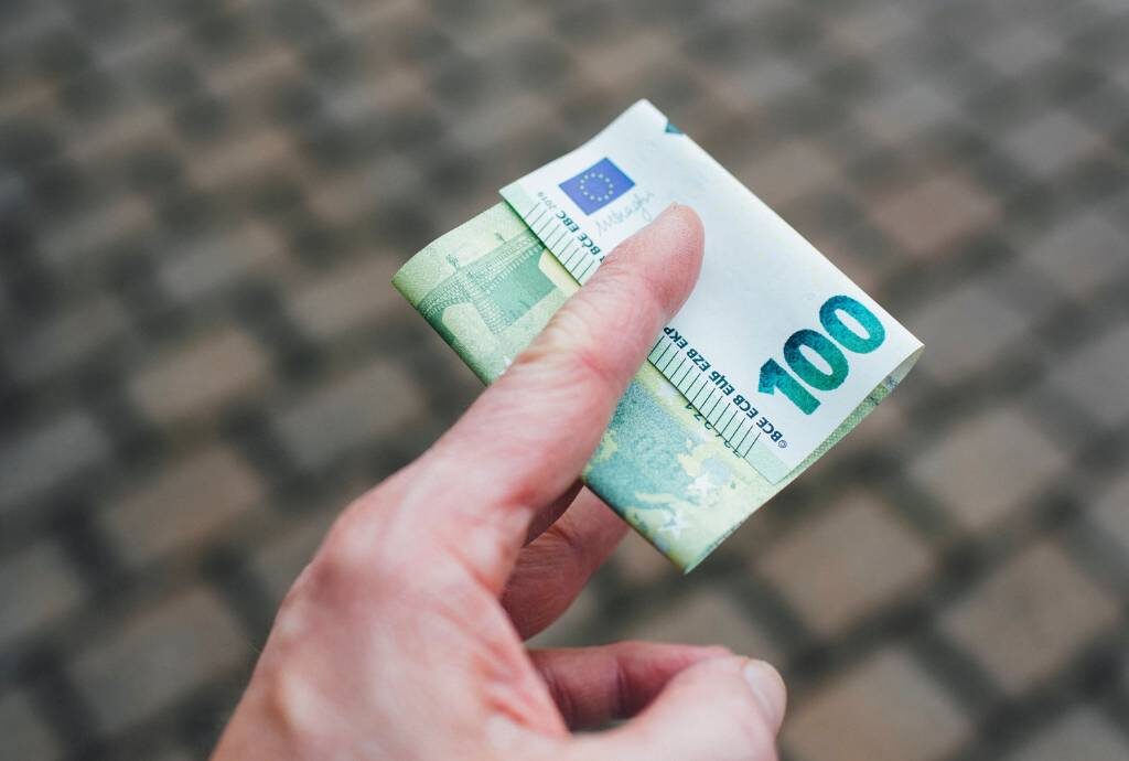 Das Bild zeigt eine Hand - nur halb im Bild und von oben betrachtet. Zwischen Zeigefinger und Mittelfinger klemmt ein 100-Euro-Schein. Im Hintergrund ist Straßenpflaster zu sehen.