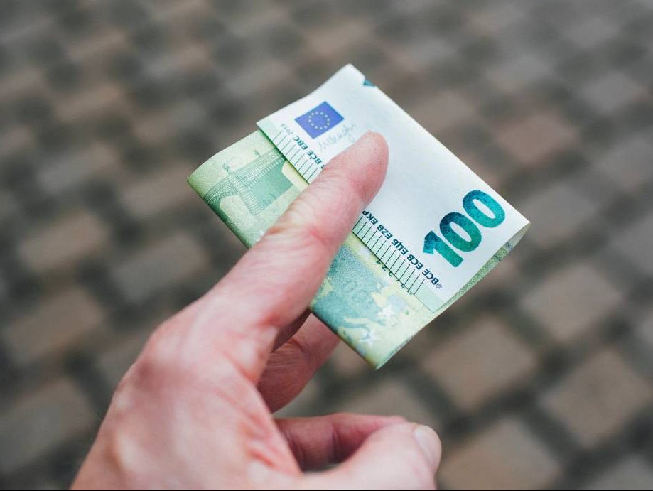 Das Bild zeigt eine Hand - nur halb im Bild und von oben betrachtet. Zwischen Zeigefinger und Mittelfinger klemmt ein 100-Euro-Schein. Im Hintergrund ist Straßenpflaster zu sehen.