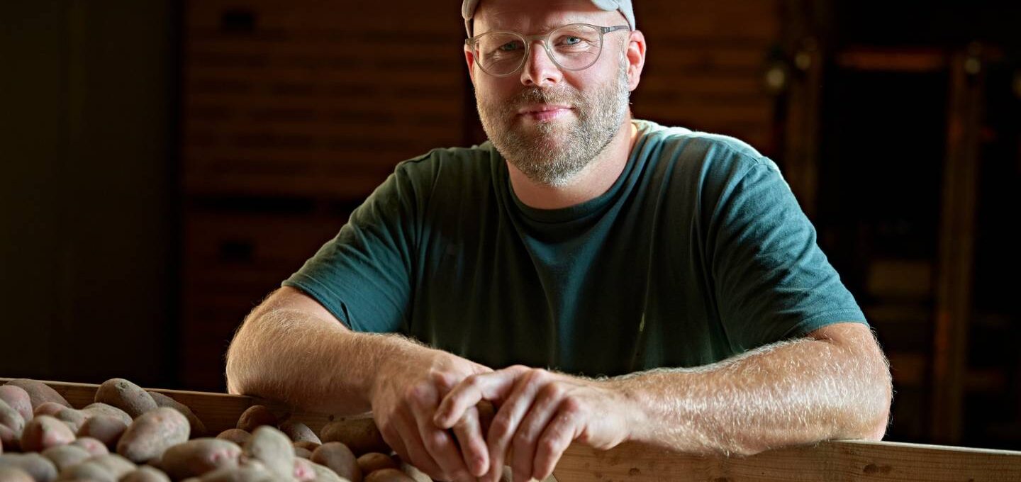 Bio-Landwirt Johann Gerdes stützt sich mit den Unterarmen auf einer Kiste Kartoffeln ab. Er trägt Basecap, Brille und ein dunkelgrünes T-Shirt
