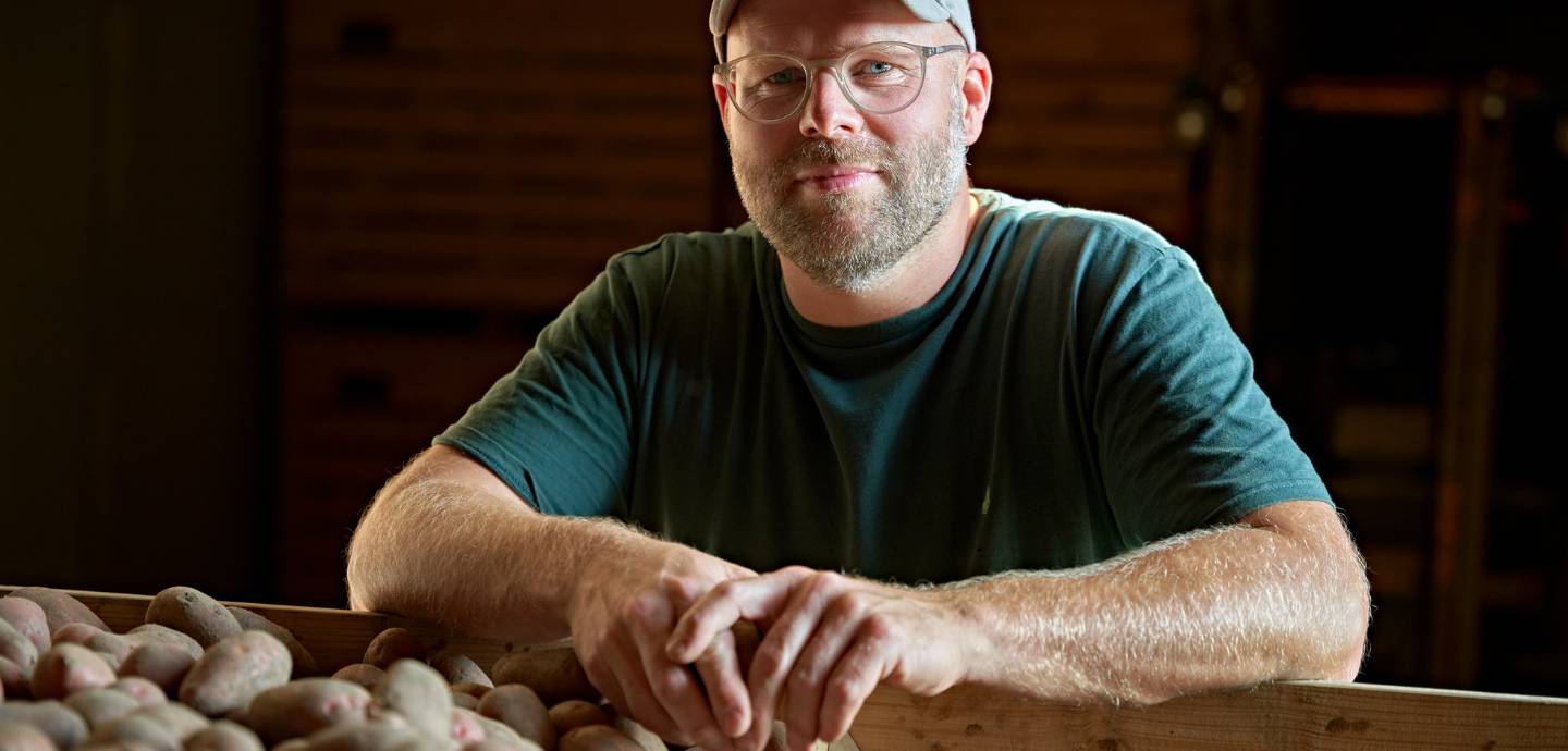 Bio-Landwirt Johann Gerdes stützt sich mit den Unterarmen auf einer Kiste Kartoffeln ab. Er trägt Basecap, Brille und ein dunkelgrünes T-Shirt