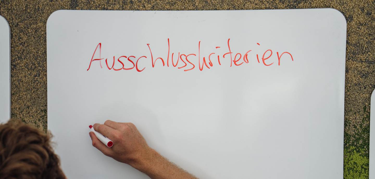 Auf einem Whiteboard steht in Handschrift das Wort Ausschlusskriterien. Darunter ist eine Hand mit Stift, die im Begriff ist zu schreiben.