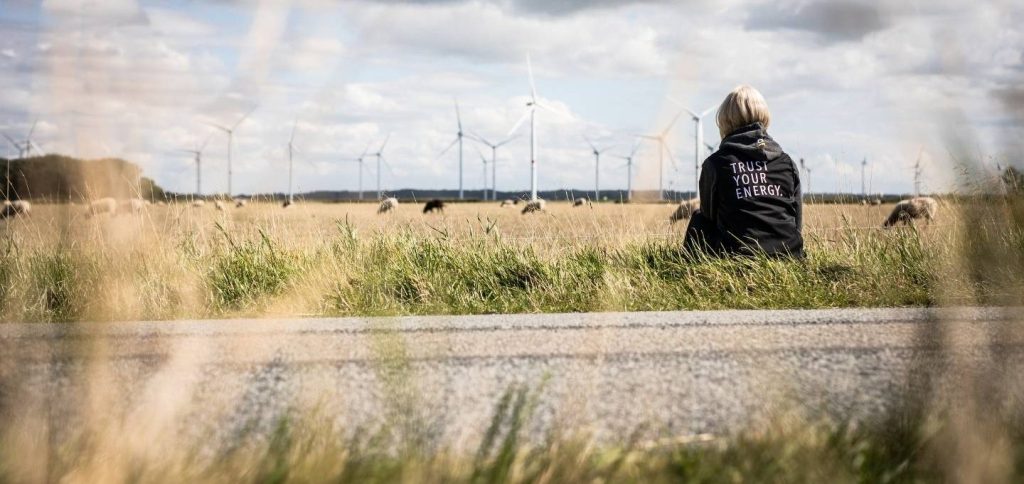 Eine Frau trägt einen Pulli mit der Beschriftung "Trust your Energy". Sie sitzt vor einer Schafwiese, im Hintergrund sind zahlreiche Windräder zu sehen.