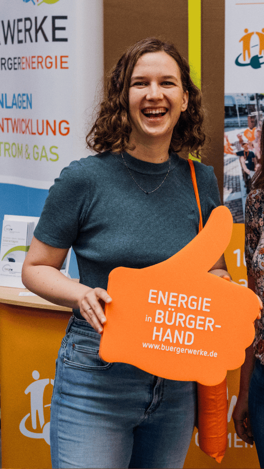 Auf dem Bild ist Sara Haug von der Bürgerenergiegenossenschaft Bürgerwerke zu sehen. Sie hält einen orangenes Schild in Form eines Daumens hoch mit dem Text: Energie in Bürgerhand. buergerwerke.de