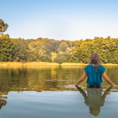 Wasserbotschafterin Anoosh Werner steht bis zur Hüfte in einem See, gelegen inmitten von Wiesen und Feldern. Sie und die Umgebung spiegeln sich im Wasser, der Himmel ist blau und leicht milchig.