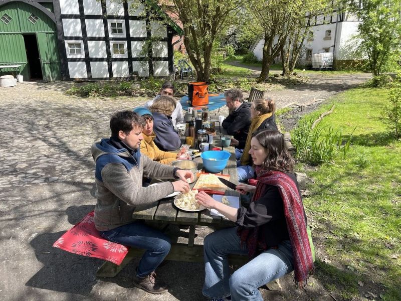 Eine Gruppe von Menschen sitzt unter freiem Himmel an einem großen Holztisch und bereitet das Gemüse zu einem gemeinsamen Kochen vor. Sie sind auf dem Hof eines alten Bauernhofs in Fachwerkstil.