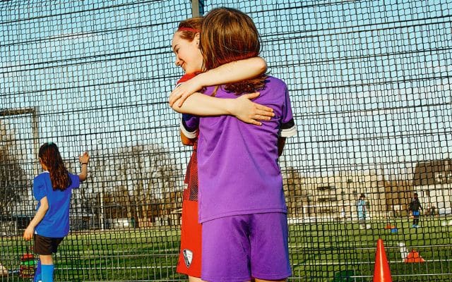 Am Rande eines sonnigen Fußballplatzes umarmen sich zwei junge Mädchen, gekleidet in farbenfrohen Fußballtrikots.