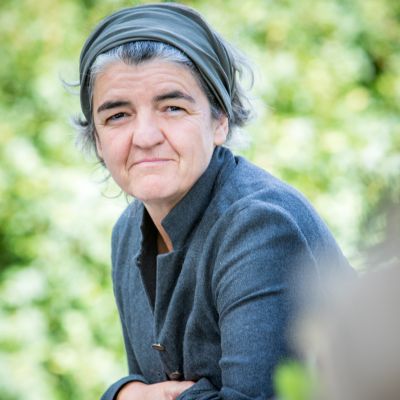 Ulrike Kreuer in schwarzem Oberteil und mit schwarzem Stirnband schaut leicht nach vorne gelehnt und mit verschränkten Armen in die Kamera. Hintergrund ist blättrig-grün